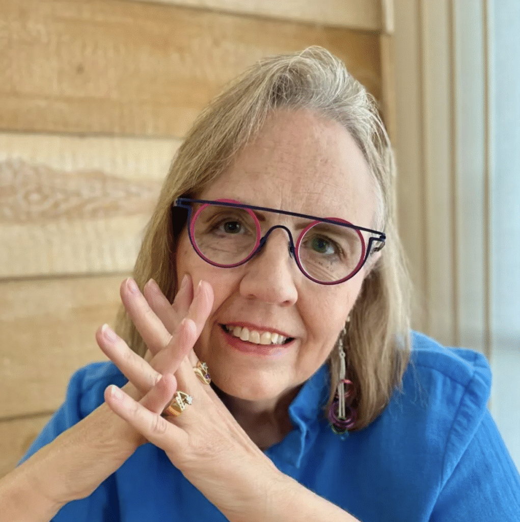 A smiling woman with light hair and colorful glasses rests her hands near her face. She is wearing a bright blue shirt, dangling earrings, and several rings, seated in front of a wooden wall with natural light coming from a nearby window.